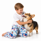 A young boy in a white t-shirt and blue patterned pajama pants hugs a corgi dog on a seamless white studio background.