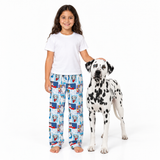 A young girl with long dark hair stands barefoot next to a Dalmatian dog on a white studio background. The girl is wearing a plain white T-shirt and blue pajama pants with a cartoon dog print, while the dog wears a white coat with black spots.
