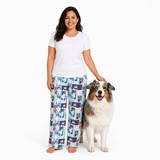 A woman wearing a white t-shirt and blue patterned pajama pants stands barefoot next to a fluffy Australian Shepherd dog on a solid white background.