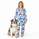 A young girl stands barefoot next to a dog wearing a matching pajama set. The pajamas are light blue with a repeating pattern of cartoon penguins in red and dark blue outfits.