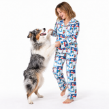 A woman wearing a blue and white cartoon-patterned pajama set stands barefoot on a white studio background while playing with a fluffy gray, white, and brown dog that is jumping up to greet her.