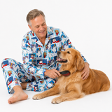 A smiling man wearing blue and white penguin-patterned pajamas sits on a white studio floor, brushing a golden retriever dog. The pajamas feature a colorful cartoon penguin print with blue, white, red, and black details.
