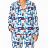 A person wearing a matching long-sleeve button-up pajama shirt and pants set with a repeating cartoon dog collage print on a light blue background. The print features white dogs, brown dogs, and red scarves against blue panels, with snowflake details.