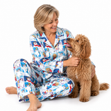 A woman wearing a blue and white cartoon-patterned pajama set sits on a white studio floor, smiling and petting a fluffy brown dog.