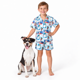 A young boy stands barefoot next to a black, white, and tan dog on a white studio background, wearing a matching short-sleeve pajama shirt and shorts set with a colorful cartoon penguin print.