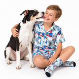 A smiling young boy in a blue cartoon pajama set with matching socks sits on the floor and hugs a black-and-white dog against a clean white background.