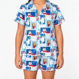 A model wearing a two-piece pajama set with a blue and white winter-themed print featuring cartoon polar bears, snowmen, and dogs. The set includes a short-sleeve button-up shirt and matching shorts.