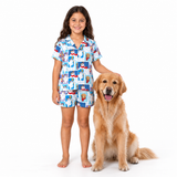 A young girl stands barefoot next to a golden retriever dog on a white studio background, wearing a matching short-sleeve pajama set with a blue base and a cartoon character print.