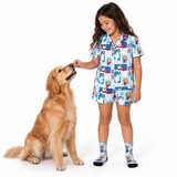 A young girl wearing a light blue cartoon-print pajama set and matching socks stands next to a golden retriever dog on a white studio background, smiling as the dog reaches up to lick her hand.