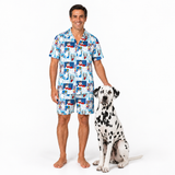 A man wearing a matching short-sleeve button-up pajama shirt and shorts set with a colorful cartoon dog print, standing next to a Dalmatian dog on a white studio background.