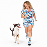 A woman wearing a matching short-sleeve button-up shirt and shorts pajama set with a cartoon dog print, walking alongside a black and white dog on a white studio background. She is also wearing matching socks.