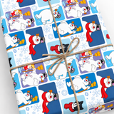 A gift-wrapped rectangular package tied with twine, featuring a winter penguin print with blue panels, white penguins, red scarves, and snowflake motifs on a light background.