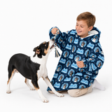 A boy kneels on a white studio floor interacting with a black, white, and tan dog. The boy is wearing a navy blue hooded blanket/sleep sack covered in a repeating light blue 'Just Dog' dog face pattern, while the dog holds a small tug toy.