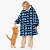 A woman wearing a navy blue hooded blanket jacket with a repeating Jack Skellington pattern, interacting with an orange tabby cat on a white background.