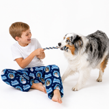 A young boy in blue and white dog-themed pajamas sits on the floor playing tug-of-war with a black and white Australian Shepherd dog on a white studio background.