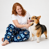 A woman with red hair sits on a white studio floor, smiling and brushing a Corgi mix dog. She is wearing navy blue pajama pants with a white and blue 'Peanuts' character pattern, a white t-shirt, and is barefoot.