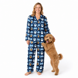 A woman wearing a navy blue pajama set with a cartoon dog pattern stands barefoot against a white studio background, holding a fluffy golden-brown dog that is standing on its hind legs.