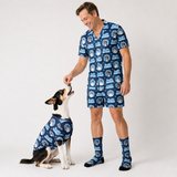 A man and a dog wearing matching navy blue patterned pajamas and socks, with the man feeding the dog, against a plain light gray studio background.