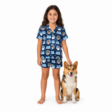 A young girl stands barefoot next to a corgi dog on a white studio background, wearing a navy blue short-sleeve pajama set with a repeating dog-face pattern and light blue accents.