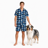 A man wearing a navy blue short-sleeve pajama shirt and matching shorts with a repeating dog print stands barefoot next to an Australian Shepherd dog on a white studio background.