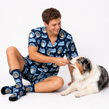A man wearing a matching navy blue pajama set with a cartoon dog print sits on a white studio floor, smiling as he feeds a fluffy Australian Shepherd dog. The set includes a short-sleeve top and shorts, with matching socks.