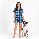 A woman stands barefoot next to a black, white, and tan dog wearing a matching blue short-sleeve pajama set with a dog-themed print. The pajamas feature a blue base with white and light blue graphics of dogs and the text 'This is my dog'.