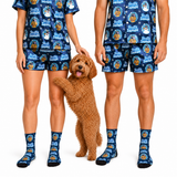 A matching family pajama set shown on two models and a dog, featuring a navy blue base with a festive snowman print and the text 'Frosty the Great'. The set includes a short-sleeve top and shorts for humans, along with matching crew socks, with a small brown poodle standing between them.