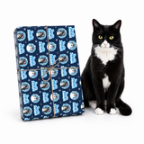 A black and white cat sits next to a wrapped gift box on a clean white background. The gift box is wrapped in navy blue paper with a repeating pattern of cartoon cats in circular frames and is tied with twine.