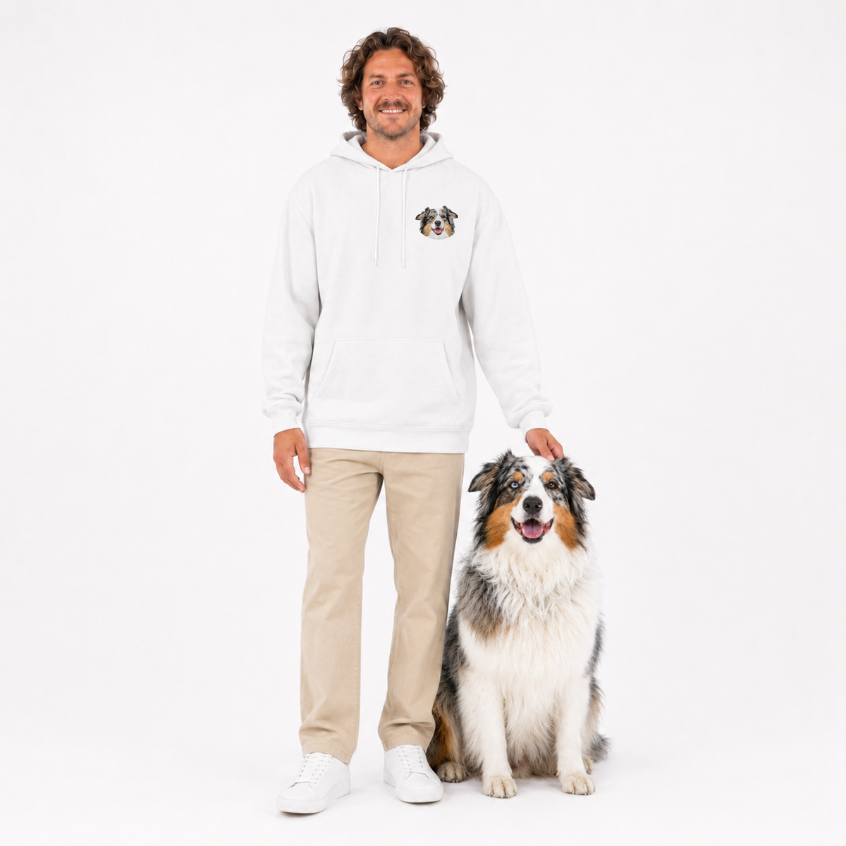 A man wearing a white pullover hoodie with a small dog graphic on the left chest, paired with beige pants and white sneakers, standing next to a fluffy Australian Shepherd dog on a solid white background.