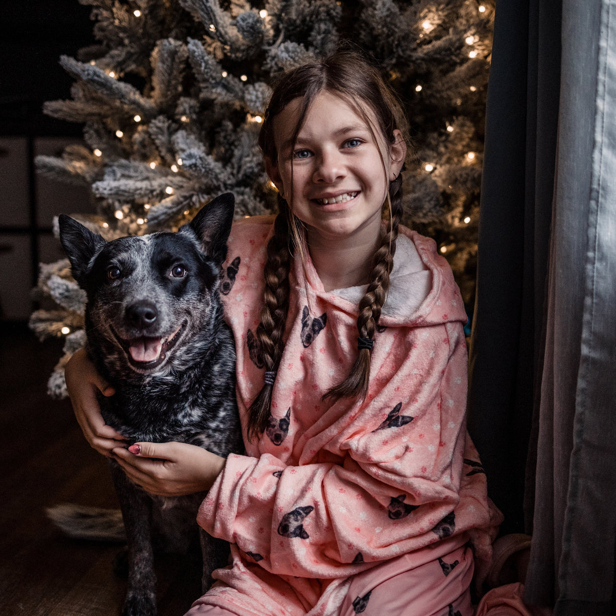 Child wearing a Light Pink Paws hooded blanket hugging a black and gray dog in front of a lit Christmas tree.
