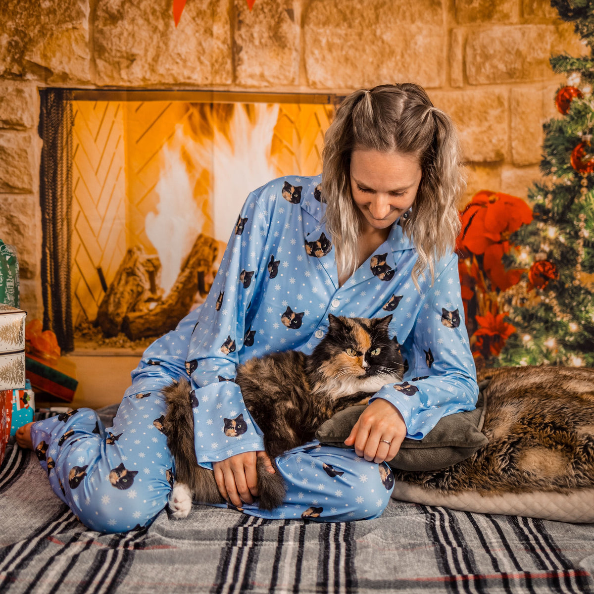 A woman with blonde hair, wearing a long-sleeve light blue pajama set patterned with cat faces and snowflakes, is sitting on a striped blanket. She is gently holding a fluffy calico cat resting on her lap.