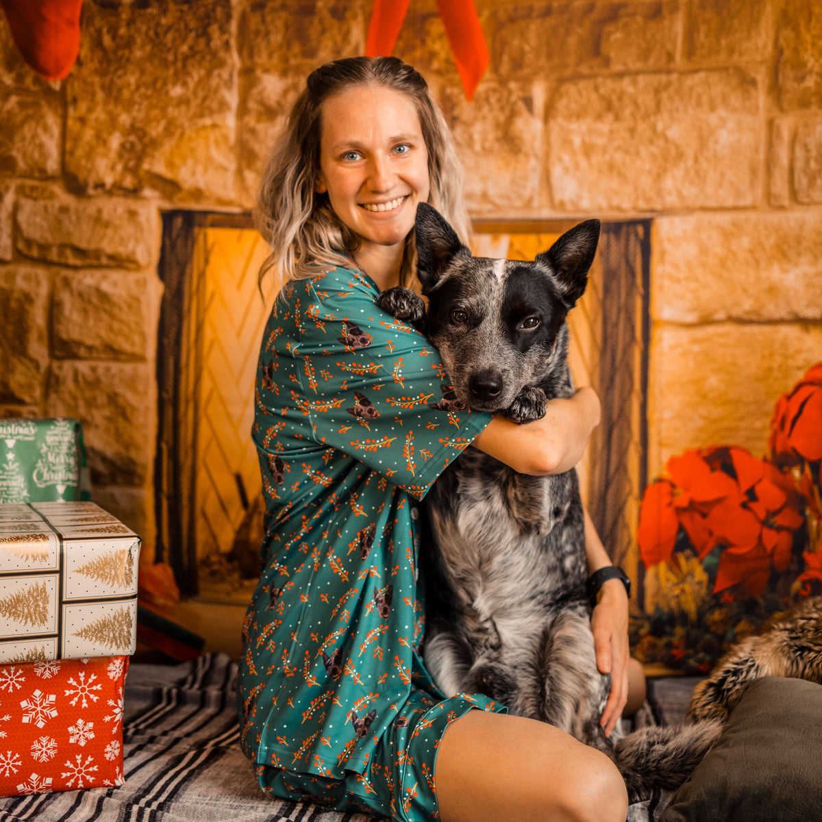 A smiling woman with blonde hair, wearing a short-sleeve teal patterned pajama set, is sitting on a blanket and hugging a black and white mottled Australian Cattle Dog.
