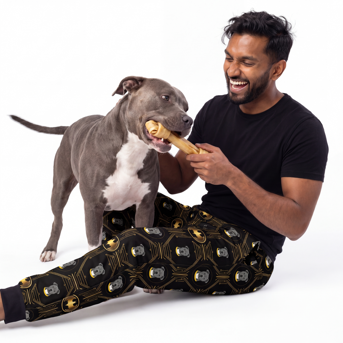 Man and dog playing with a bone wearing batman sweatpants on a white background