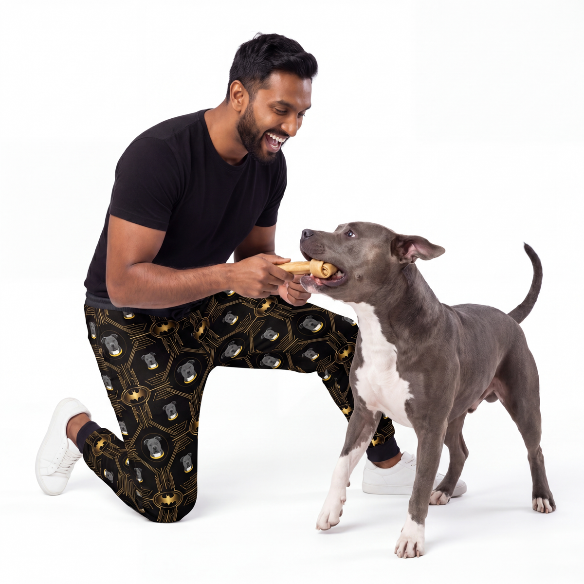 Man wearing batman sweatpants feeding a dog a treat on a white background