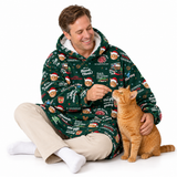 A man wearing a green Christmas-themed hooded blanket sleeper sits on the floor with a ginger cat. The sleeper features a festive print with phrases like 'DAD, come home fast' and holiday illustrations.