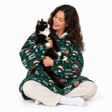 A woman sitting on a white studio floor wearing a green Christmas-themed fleece pajama robe with a black and white cat, while wearing beige pants and white socks.