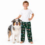 A smiling boy stands barefoot next to an Australian Shepherd dog on a white studio background, wearing dark green Christmas-themed pajama pants with holiday illustrations and a plain white T-shirt.