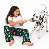A young girl with long brown hair sits on a white studio floor, playfully pulling a Dalmatian dog with a rope toy. She is wearing dark green Christmas-themed pajama pants with a white t-shirt, against a clean white background.