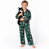 A smiling young boy stands barefoot against a white studio background, holding a black-and-white cat. He is wearing a long-sleeve pajama set with a dark green Christmas print featuring holiday illustrations and text.