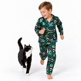 A young boy wearing a green Christmas-themed pajama set with a cat walking beside him on a clean white studio background. The pajamas feature a festive print with Santa, reindeer, snow globes, and holiday text.