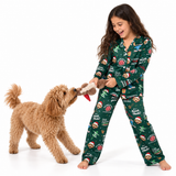 A young girl with long brown hair smiles while playing tug-of-war with a fluffy tan dog. She is wearing a dark green Christmas-themed pajama set with a festive print of Santa Claus, reindeer, gingerbread cookies, and holiday phrases like 'Merry Christmas' and 'D.