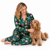 A woman wearing a green Christmas-themed pajama set sits on the floor next to a fluffy golden-brown dog, smiling and petting it. The pajamas feature a festive print with dogs, Santa hats, trees, and holiday text like 'A Great Christmas Day' and 'Merry Christmas.