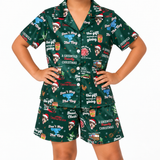A child modeling a matching short-sleeve button-up pajama shirt and shorts set in dark green with a festive Christmas print featuring the Trog dog character, Santa hats, gifts, and holiday text.