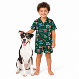 A young boy wearing a green Christmas-themed pajama set stands next to a black and white dog on a white studio background. The pajama set features a short-sleeve button-up shirt and matching shorts with a festive holiday print including Santa Claus, Christmas trees, and holiday quotes.