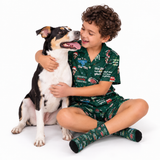 A smiling boy with curly brown hair sits on the floor hugging a black, white, and tan dog. The boy is wearing a dark green Christmas-themed pajama set with matching socks, featuring festive illustrations and text like 'The Tree of Christmas' and 'Green Wolf'.