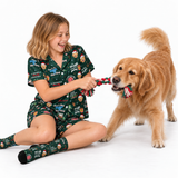 A young girl with blonde hair sits on a white studio floor, smiling and playing with a golden retriever dog. The girl is wearing a dark green pajama set with a colorful cartoon dog print, and the dog is holding a red and green tug-of-war rope toy.
