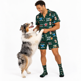 A man and his dog wearing matching dark green cartoon-print pajama set and socks, standing on a white studio background. The man is smiling and petting the dog, which is standing on its hind legs.