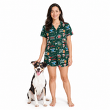 A woman stands barefoot next to a black, white, and tan dog on a white studio background, wearing a dark green short-sleeve pajama set with a colorful printed patchwork pattern featuring pizza, burger, and coffee graphics.