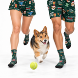 A corgi dog runs toward a yellow tennis ball on a white studio floor while two people in matching dark green Christmas-themed socks and shorts jog past, all featuring a festive print with text, dogs, and holiday motifs.