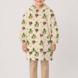 A child wearing a cream-colored hooded sweatshirt with an all-over festive print of Santa-clad elves, candy canes, snowflakes, and dog faces. The child is also wearing beige pants, standing against a plain light gray studio background.